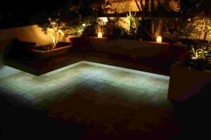 Outdoor patio with an L-shaped cushioned bench, surrounded by plants and softly illuminated by candles, accent lights, and thoughtfully placed outdoor lighting for a cozy nighttime ambiance.