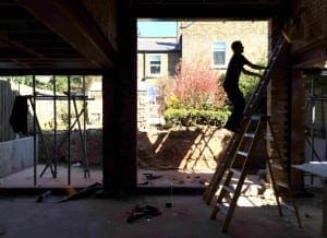 A person stands on a ladder inside a partially constructed building in Queens Park, with an open wall revealing a garden and brick houses outside.
