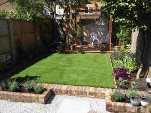 A small backyard garden with a rectangular fake lawn, brick edging, flower beds, and a wooden pergola with outdoor seating in the background.