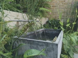 A bespoke garden feature, this rectangular stone water trough with a metal spout is surrounded by lush green plants and ferns against a brick wall background.