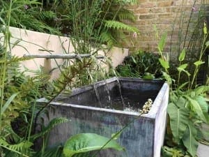 A rectangular metal water trough, one of the garden’s striking water features, with water flowing from two pipes, is surrounded by green plants and ferns, set against a brick wall and lush garden backdrop.