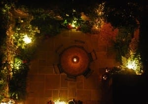 A round table with four chairs is centered on a stone patio, surrounded by plants and enhanced by warm outdoor lighting that creates an inviting ambiance at night.