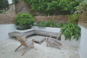Small modern patio with wooden chairs, a floating bench, tile flooring, and raised planters filled with green plants against slatted wooden privacy fences.