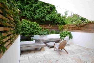 Modern patio with a wooden lounge chair, low table, floating bench seating, potted plants, tiled floor, and wooden privacy fencing surrounded by lush greenery.