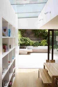 A modern dining area with a wooden table, chairs, and a floating bench beside a white bookshelf, featuring a glass ceiling and sliding doors opening to a bright garden patio.