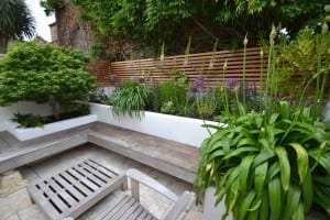 A modern garden patio featuring a floating bench, potted plants, a slatted wooden fence, and a raised flower bed filled with lush greenery and purple flowers.