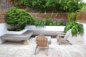 A small patio with light stone tiles features a floating bench, two wooden chairs, potted plants, and a raised planting bed with lush greenery and flowers, all bordered by wooden fencing.