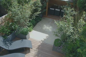 Modern backyard with wooden deck, two black lounge chairs, potted plants, a tiled patio area, and a shaded outdoor room featuring dark furniture in the background.