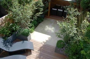 A small landscaped patio serves as a stylish outdoor room, featuring wooden decking, two black lounge chairs, potted plants, and a covered seating area with chic black and white furniture.