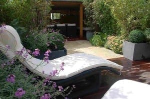 A modern garden patio featuring a white cushioned lounge chair, flowering plants, potted shrubs, and a shaded seating area in the background creates an inviting outdoor room perfect for relaxation.