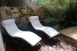 Two white cushioned lounge chairs sit on a wooden deck in a small garden, creating an inviting outdoor room with potted plants and a wooden fence covered in climbing greenery.