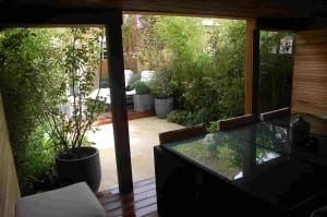 Covered patio designed as an inviting outdoor room, featuring a glass table and chairs overlooking a small garden area with potted plants and cozy seating.
