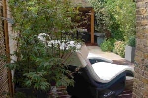 A cushioned lounge chair sits on a wooden deck in an inviting Outdoor Room, surrounded by green plants and brick walls in a private garden.