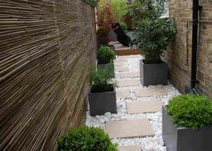 Narrow garden path with square stepping stones, white decorative rocks, potted plants, and a bamboo fence on one side—perfect for an outdoor room retreat—nestled beside a brick wall.