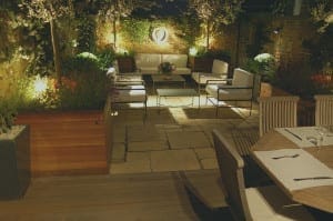 Outdoor patio with stone flooring, surrounded by plants and trees, features cushioned seating, a coffee table, and a wooden dining table set under evening lighting—creating a cozy Night Garden ambiance.
