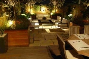 A modern outdoor patio at night features cushioned seating, a dining table, potted plants, and ambient lighting against brick walls—perfect for relaxing in your own Night Garden retreat.