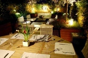 A well-lit Night Garden backyard patio at night features a wooden dining table set for six in the foreground, with a cozy seating area of sofas and plants in the background.