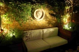 Outdoor seating area with a white cushioned bench, brick wall, lush greenery, and soft lighting creates a relaxing Night Garden vibe; a decorative round plaque hangs on the wall.