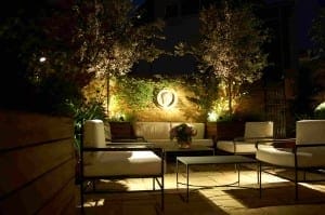 Outdoor patio at night with cushioned chairs, a coffee table, potted plants, and warm lighting illuminating a brick wall and trees creates an inviting Night Garden ambiance.