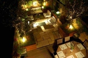A modern outdoor patio at night, transformed into a Night Garden with illuminated garden lights, white seating, a dining table set for six, and potted plants decorating the brick walls.