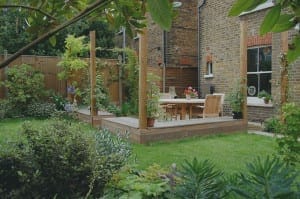 A wooden deck with a table and chairs is set in a garden with green grass, edible plants, and a brick house in the background.