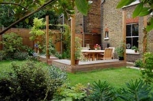 A wooden patio with a table and chairs sits in a green garden next to a brick house, surrounded by edible plants and a fenced yard.