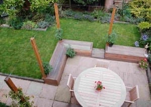 Aerial view of a garden with a patio area, round table and chairs, wooden benches with potted edible plants, and a lawn bordered by flower beds.