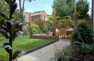 A landscaped garden with a paved path, green lawn, raised wooden deck, outdoor dining table with chairs, and various plants and flowers, including edible plants for a touch of fresh flavor right from your backyard.