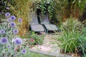Two empty lounge chairs are placed side by side on a stone patio, surrounded by dense green foliage, blooming flowers, and a selection of edible plants in the garden.