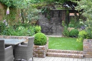 A small garden with a patio area, outdoor table and chairs, manicured lawn, brick walls, and a secluded seating area under a metal pergola with stylish pergolas beams.