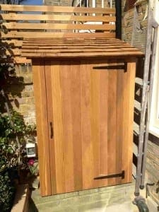 A small wooden binshed with a sloped roof and metal hinges is positioned against a brick wall in an outdoor area.