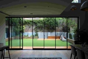 View from inside a modern home looking out through large glass sliding doors onto a backyard with fake lawns, wooden benches, and a small soccer goal.
