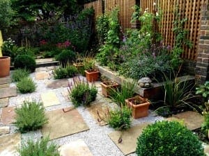 A small garden with stone pavers, gravel paths, potted plants, flowering shrubs, and a wooden trellis against a brick wall—perfect for Edible Gardens enthusiasts seeking style and function.