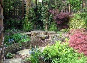 A cozy garden space with woven fences, stone paths, tree stump seats, and a variety of green and red-leaved plants under trellises—perfect for those who love tranquil gardens.