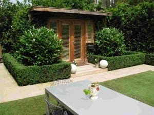 A small wooden garden shed with glass doors creates a cozy outdoor room, surrounded by manicured hedges and shrubs, with a patio table and chairs on the lawn in the foreground.