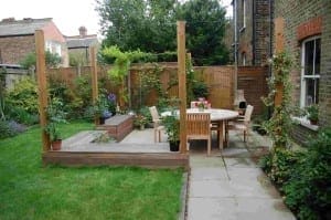 A small backyard patio with a wooden deck, round table and chairs, surrounded by plants and lawn within a wooden fence, creates a bespoke garden retreat beside the brick house.