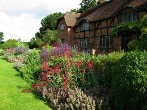 A traditional brick house with timber framing stands behind a lush garden buzzing with bees among various colorful flowers and green plants under a partly cloudy sky.