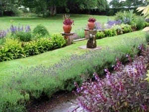 A well-maintained garden with flowering plants that attract bees, a green lawn, two large flower pots on stone pedestals, and trees in the background.