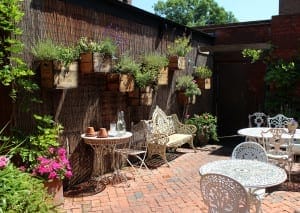 Outdoor patio with white wrought iron tables and chairs, a decorative bench, potted plants attracting bees, and wall-mounted wooden planters on a brick surface.