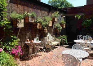 Outdoor patio with wrought iron tables and chairs, brick flooring, potted plants, and crates mounted on the fence used as herb planters.