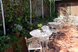 Outdoor patio with white wrought iron tables and chairs on a brick floor, surrounded by green plants, fragrant herbs, and flowers along a wall. Sunlight casts gentle shadows across the area.