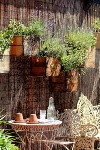 A wrought iron table and chair set sits by a wall with wooden crate planters full of fragrant herbs and flowers; a glass bottle and cups rest on the table.