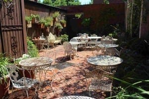 Outdoor patio area with metal tables and chairs on a brick floor, surrounded by potted plants and a wooden fence adorned with wall-mounted herb planters.