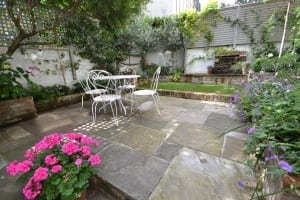 A stone patio with a shabby chic white metal table and chairs, surrounded by green plants, flowers, and a small brick water feature set against a white fence.