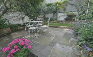 A small patio garden with stone paving features shabby chic charm—a white metal table and chairs, pink and purple flowers, and lush green plants lining the edges.