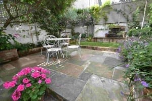 A small garden patio with stone paving, shabby chic white metal table and chairs, surrounded by green plants and pink flowers, bordered by a white fence.