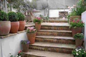 Stone steps with potted flowers lead to a shabby chic patio area, featuring a round metal table and chairs, all surrounded by greenery and wooden fencing.