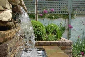 A close-up of a shabby chic stone fountain with water flowing, next to a flower bed of green plants and purple allium flowers, set against a brick wall and trellis in a charming garden.