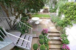A small, paved garden with a shabby chic white round table and chairs, potted plants, flower beds, and metal stairs leading down to the patio area.