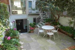 A small patio garden with stone paving, lush green plants, and a shabby chic white metal table with three chairs under a tree. Steps lead up to a glass door and windows on the house.